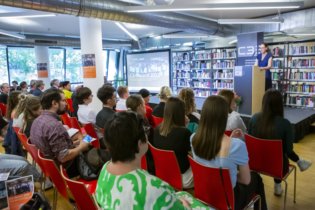 Veranstaltung: eine Person steht auf Bühne hinter Rednerpult und spricht, viele Menschen sitzen im Publikum