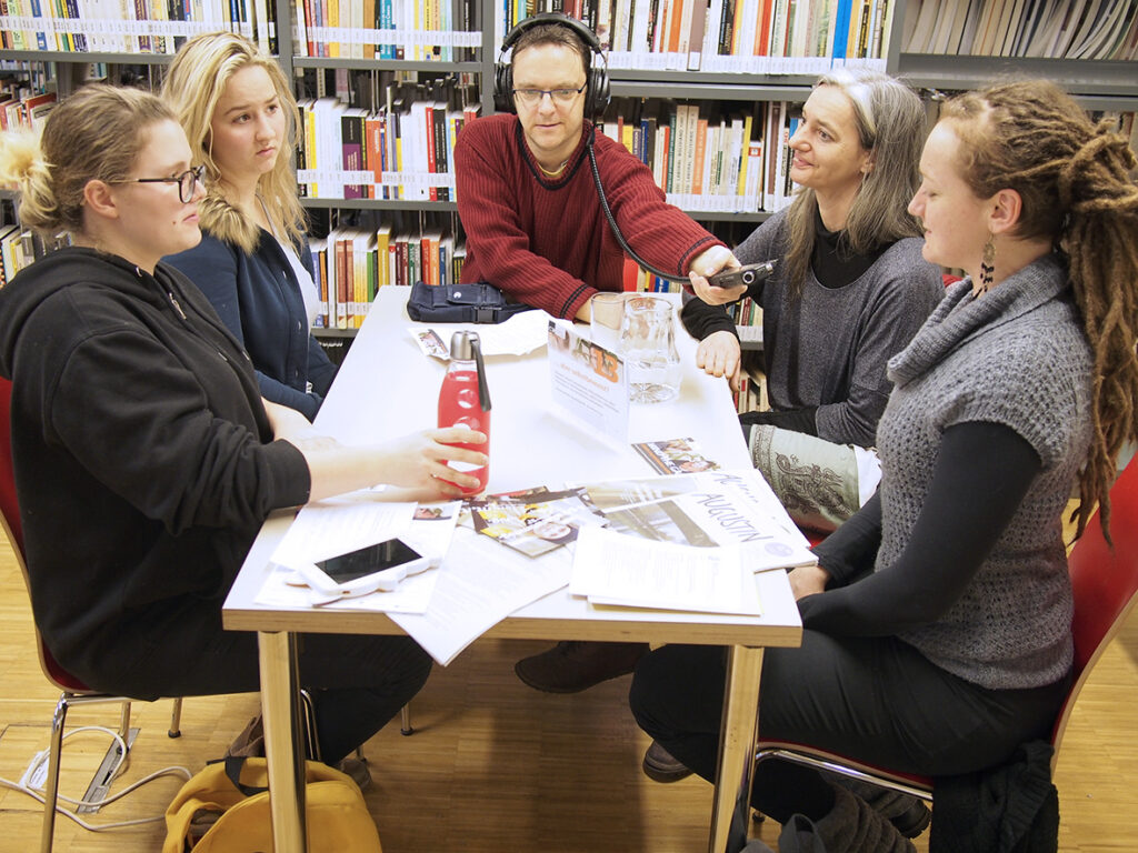 Personen bei einem Interview fürs Radio. Sie sitzen gemeinsam an einem Tisch vor einem Bücherregal