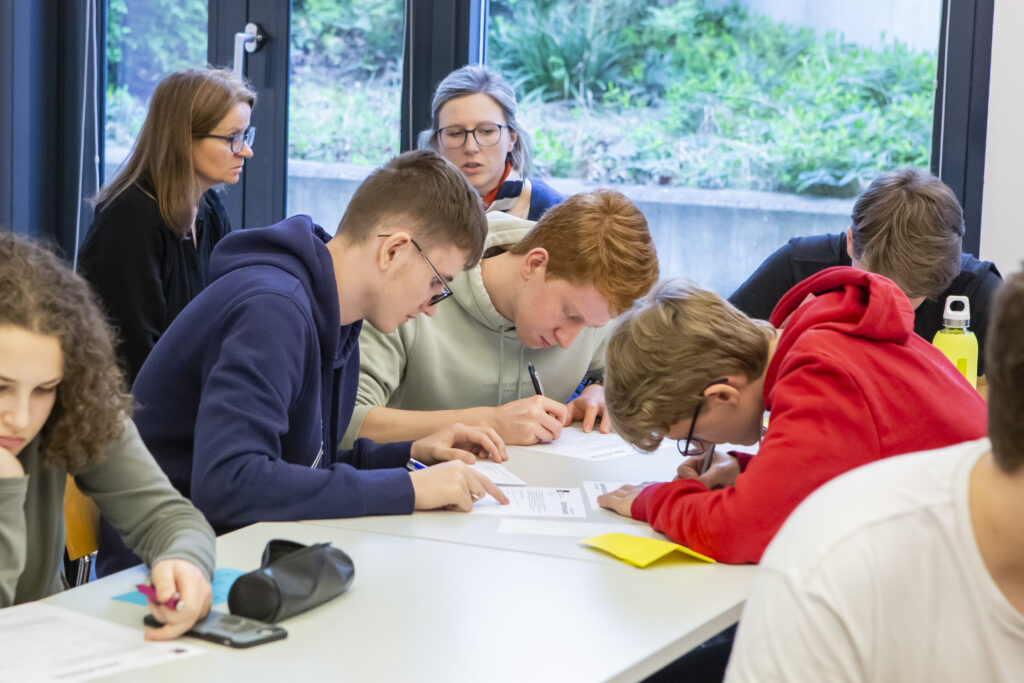 zu sehen ist eine Gruppe junger Menschen in einem Seminarraum. Sie sitzen an Tischen und arbeiten schriftlich etwas aus.
