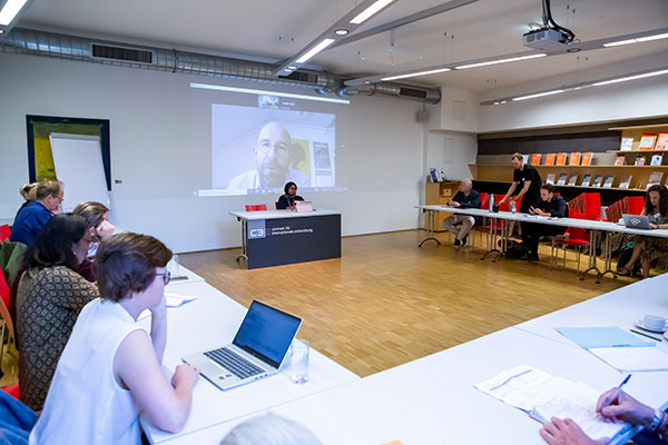 hybride Veranstaltung mit Menschen an Tischen. Eine Person sitzt am Podium und eine Person wird per Videokonferenz zugeschaltet